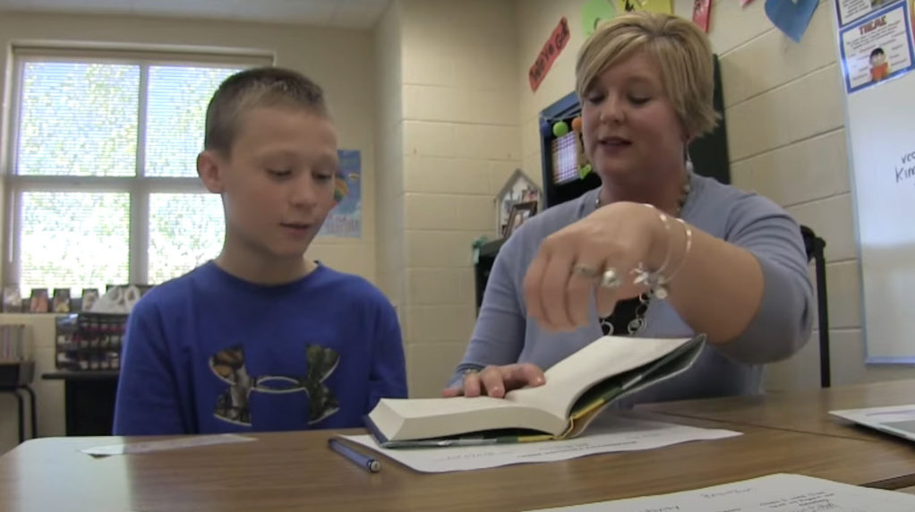 A teacher reading a book to a student at a desk in the classroom.