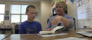 A teacher reading a book to a student at a desk in the classroom.